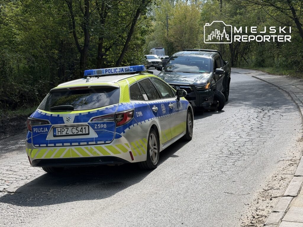 Policyjny radiowóz z niebieskimi światłami stoi na drodze, obok uszkodzony pojazd terenowy z przebitą oponą.