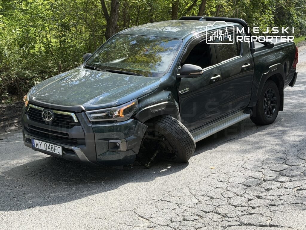 Zielony pickup z uszkodzoną przednią oponą stoi na nierównej drodze w otoczeniu drzew.
