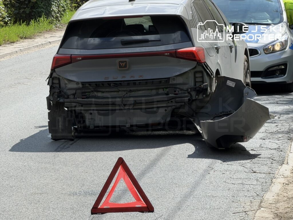 Uszkodzony samochód z odczepionym zderzakiem stoi na drodze, obok trójkąt ostrzegawczy. W tle widać inny pojazd.
