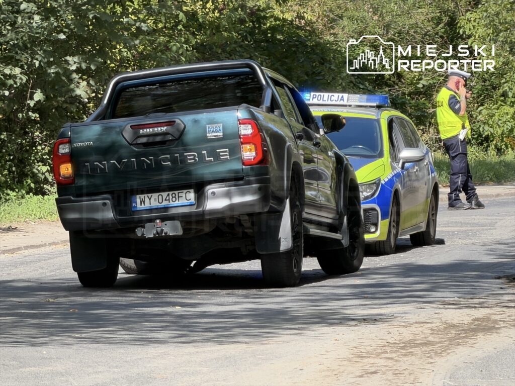 Toyota z napisem "INVINCIBLE" zatrzymana na poboczu, obok stoi radiowóz policji, a funkcjonariusz rozmawia przez telefon.