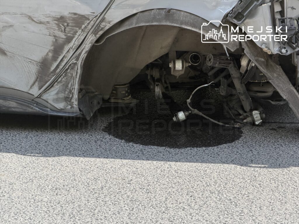 Uszkodzone auto z wgniecioną karoserią leży na drodze, z widocznymi wyciekami płynów pod pojazdem.