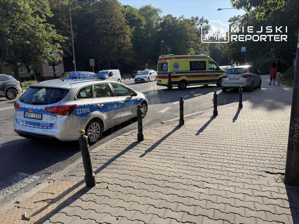 Radiowóz policyjny z niebieskimi światłami stoi na ulicy obok karetki pogotowia, w tle widać drzewa.