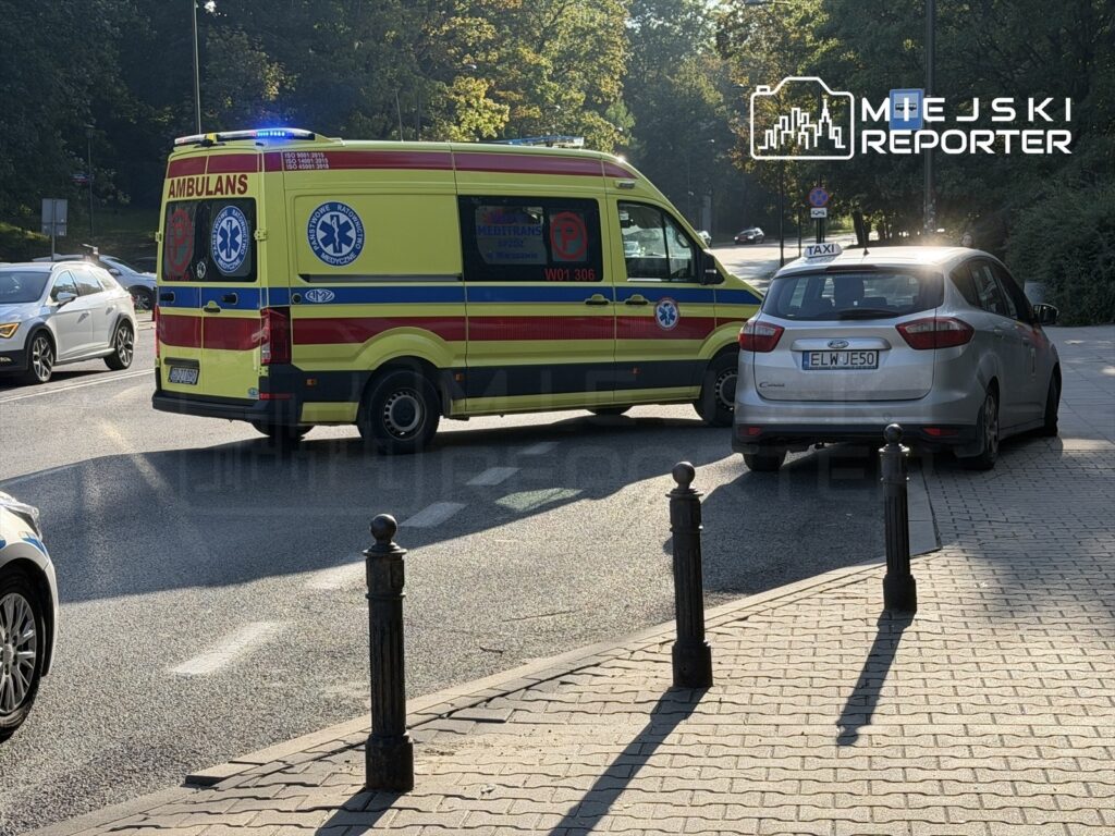 Karetka pogotowia z włączonymi sygnałami świetlnymi zatrzymana obok samochodu osobowego na ulicy.