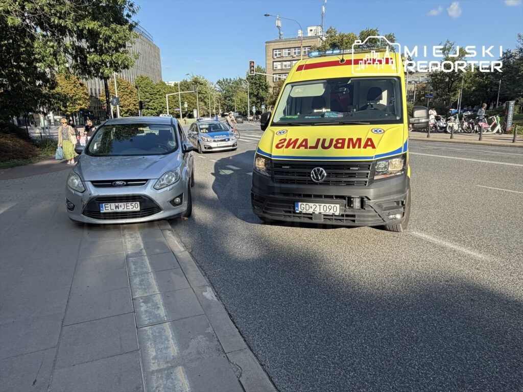 Srebrny samochód osobowy zaparkowany na chodniku obok karetki pogotowia z włączonymi sygnałami świetlnymi.