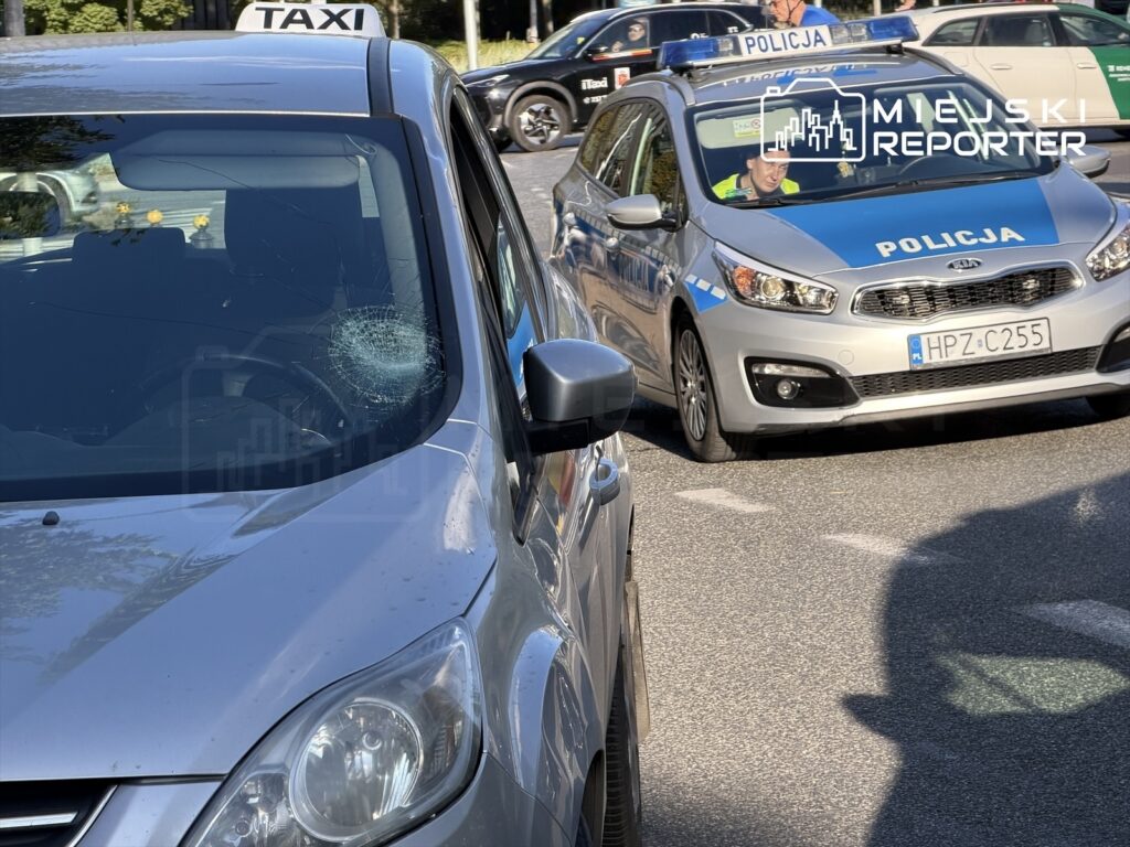 Uszkodzony samochód osobowy z pękniętą szybą stoi na drodze, obok patrolujący policjant w radiowozie.