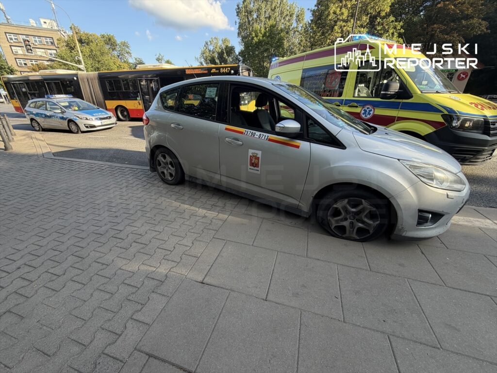 Samochód osobowy z uszkodzoną oponą zaparkowany na chodniku obok przystanku autobusowego, w tle widoczne autobusy i karetka.