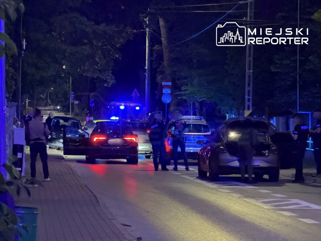 Policjanci zabezpieczają miejsce zdarzenia, podczas gdy jeden z funkcjonariuszy przeszukuje samochód na oświetlonej ulicy.