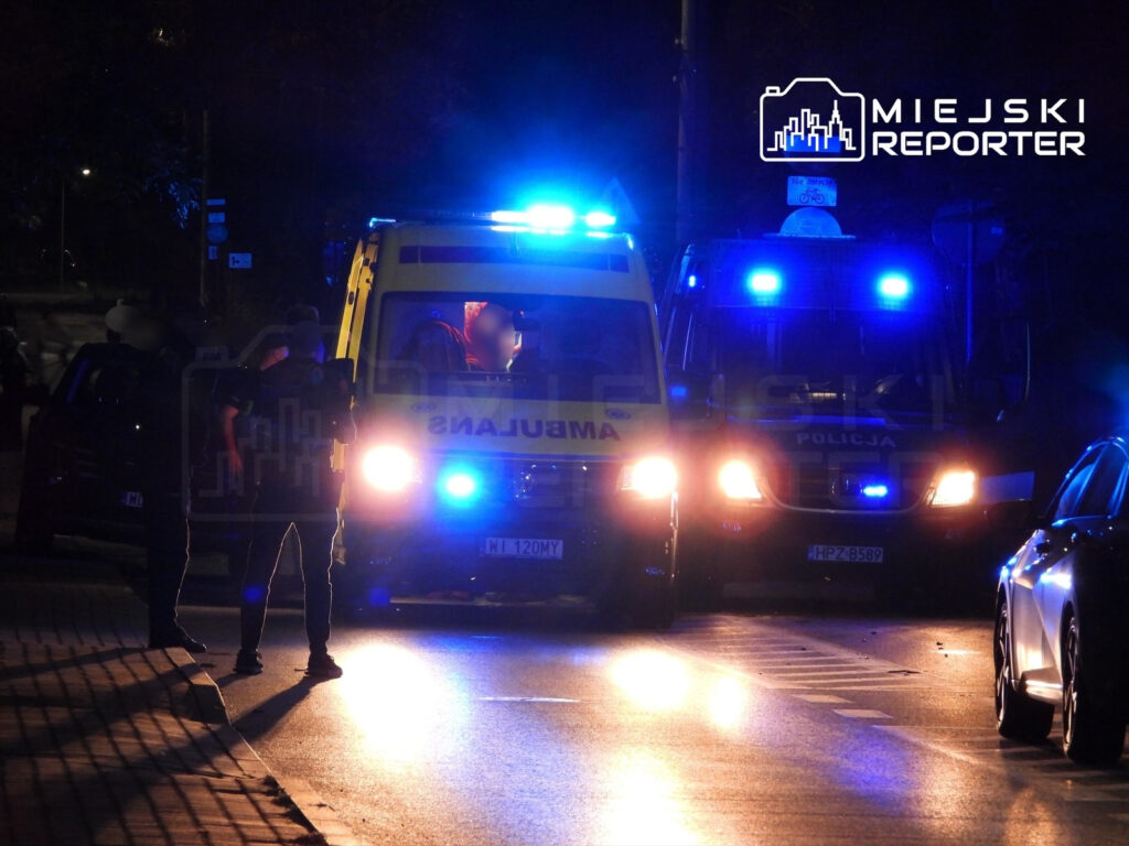 Grupa policjantów prowadzi interwencję przy zaparkowanej karetce pogotowia w nocy.
