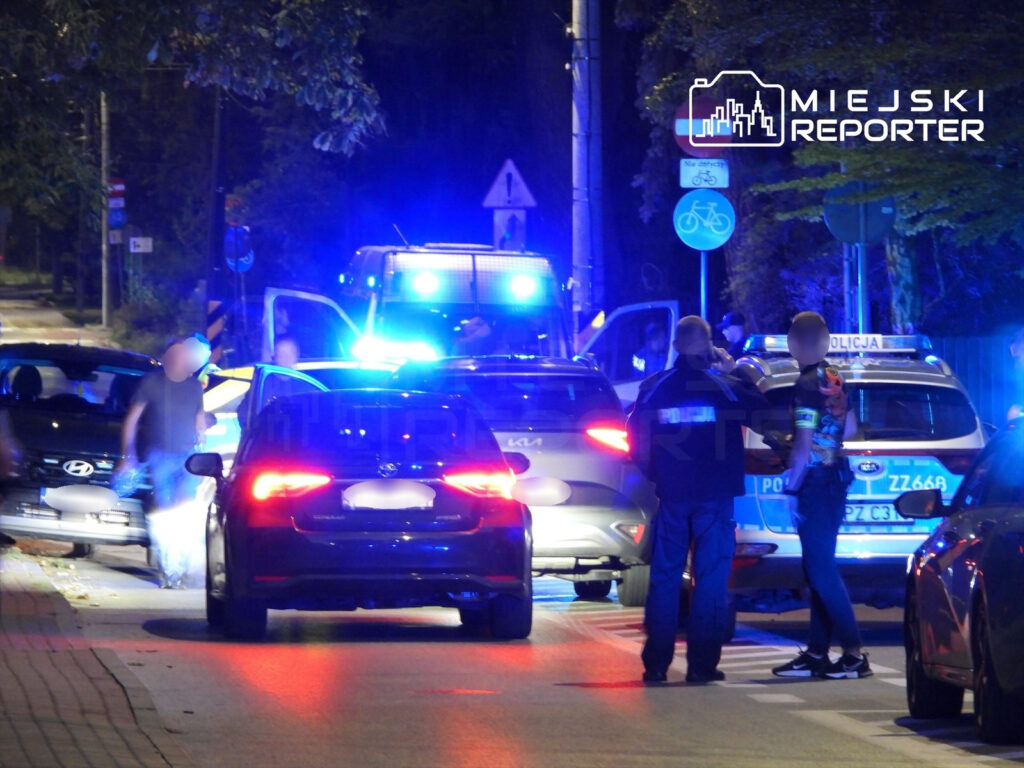 Policjanci w mundurach rozmawiają na oświetlonej ulicy, obok zatrzymanych pojazdów z włączonymi sygnałami świetlnymi.