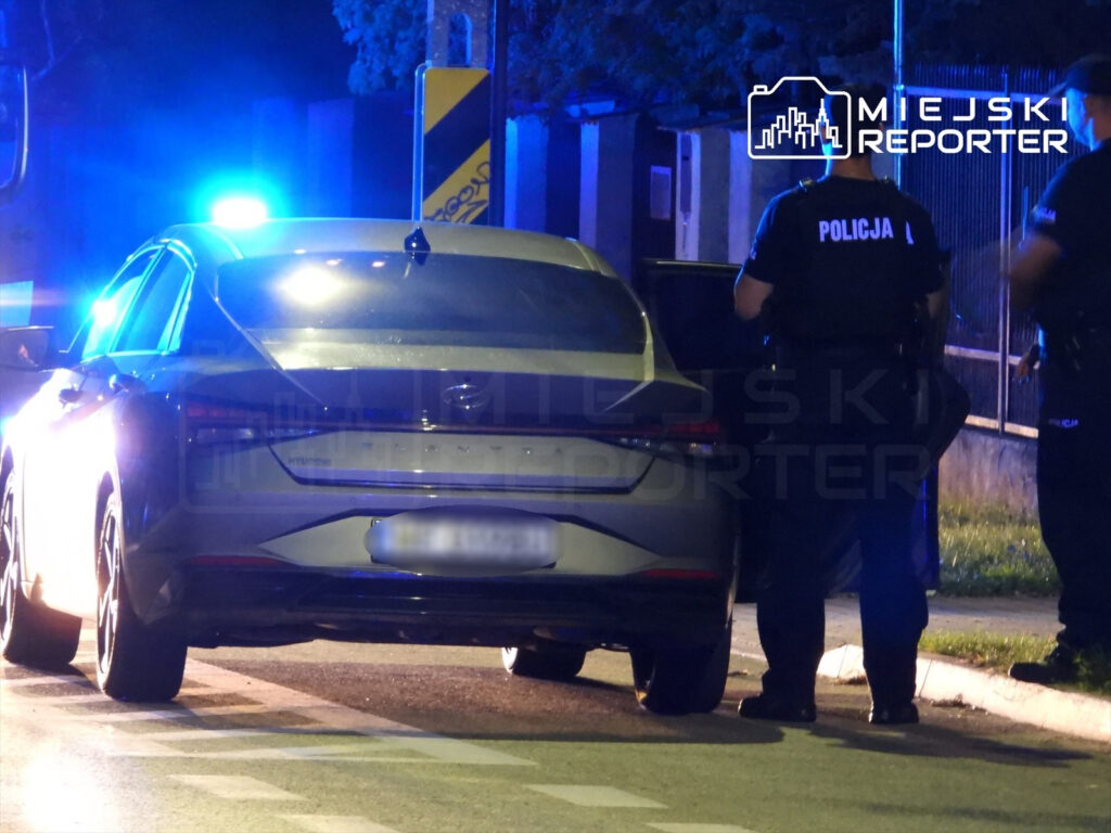 Policjanci w mundurach przeszukują samochód osobowy w nocy, w tle migające niebieskie światła radiowozu.