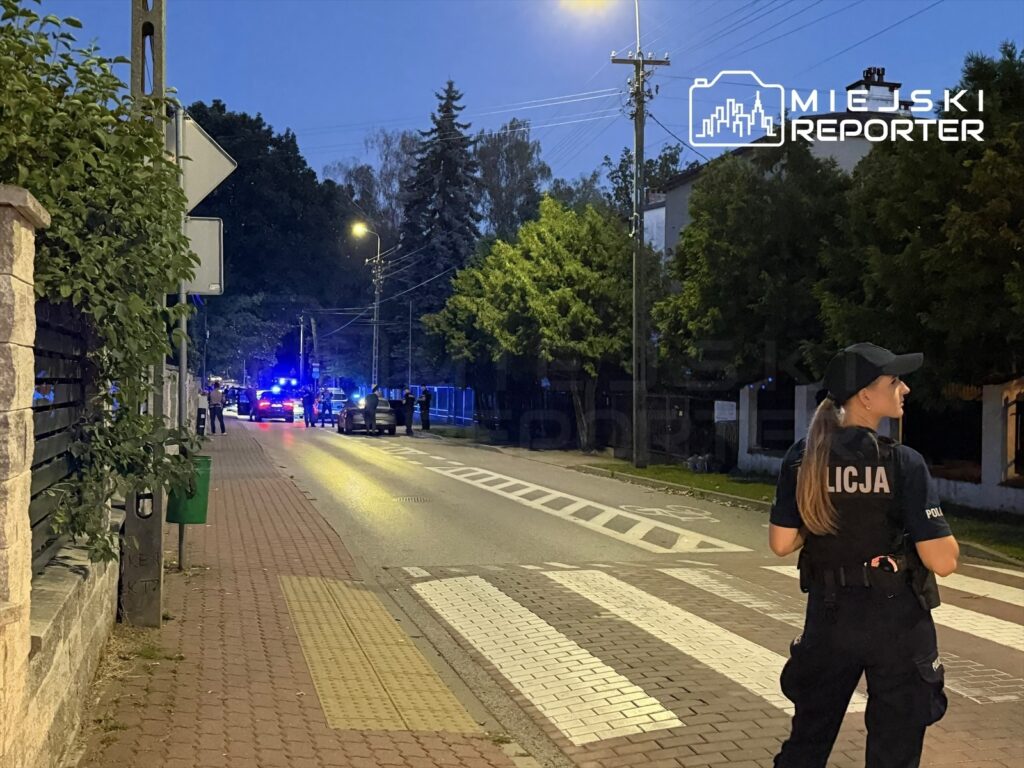 Policjantka w mundurze obserwująca sytuację na oświetlonej ulicy, w tle radiowozy z włączonymi sygnałami świetlnymi.