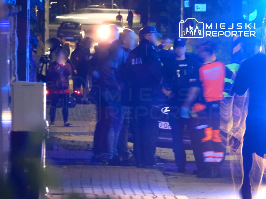 Grupa policjantów i ratowników medycznych rozmawia na ulicy, w tle widać radiowóz i pojazdy.