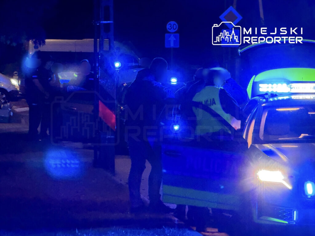 Grupa policjantów w mundurach rozmawia przy radiowozie w nocy, otoczeni niebieskimi światłami.