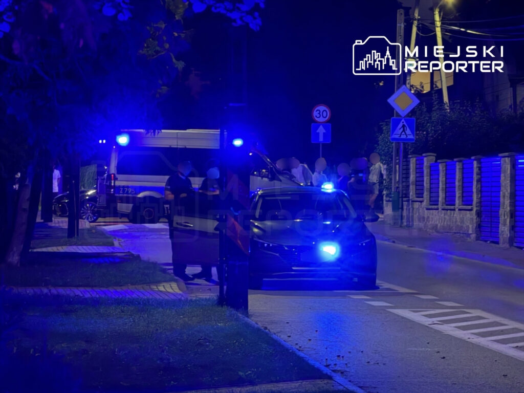 Policjanci w mundurach rozmawiają obok radiowozu z włączonymi sygnałami świetlnymi na nocnej ulicy.