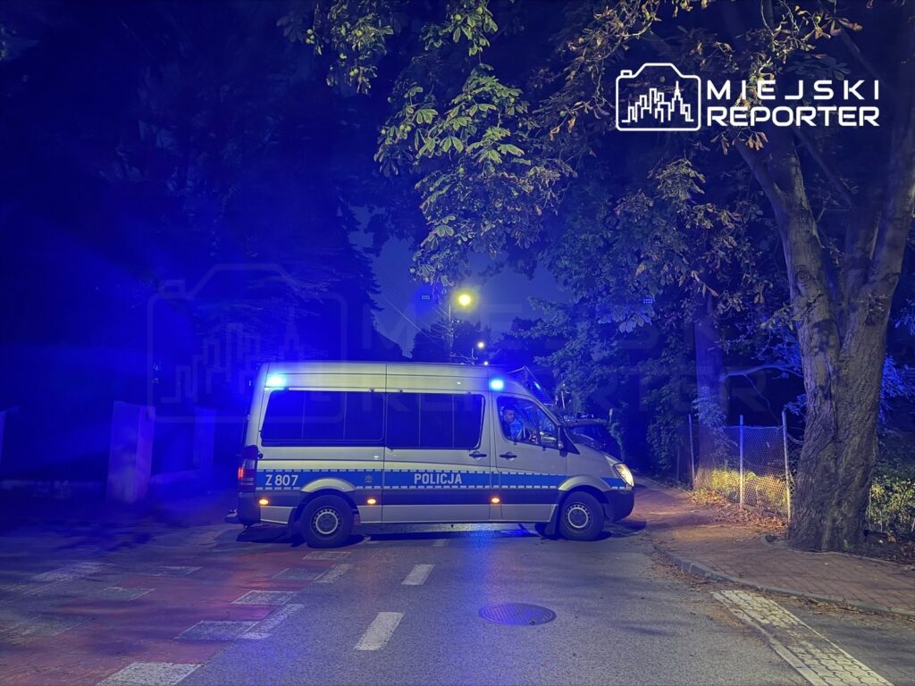 Policyjny van z niebieskimi sygnałami świetlnymi stoi na ulicy w ciemnej okolicy, otoczonej drzewami.