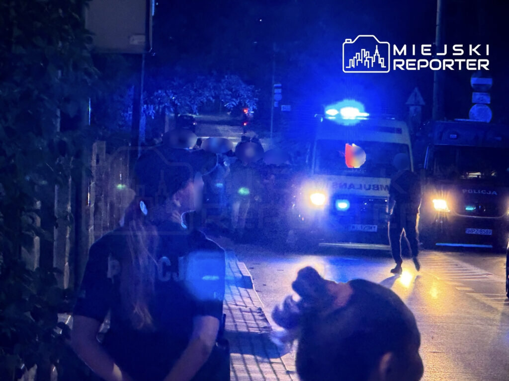 Policjantka w mundurze obserwuje działania służb ratunkowych na oświetlonej niebieskimi światłami ulicy.