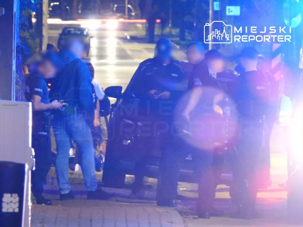 Grupa policjantów w mundurach prowadzi interwencję w nocy przy zaparkowanym samochodzie.