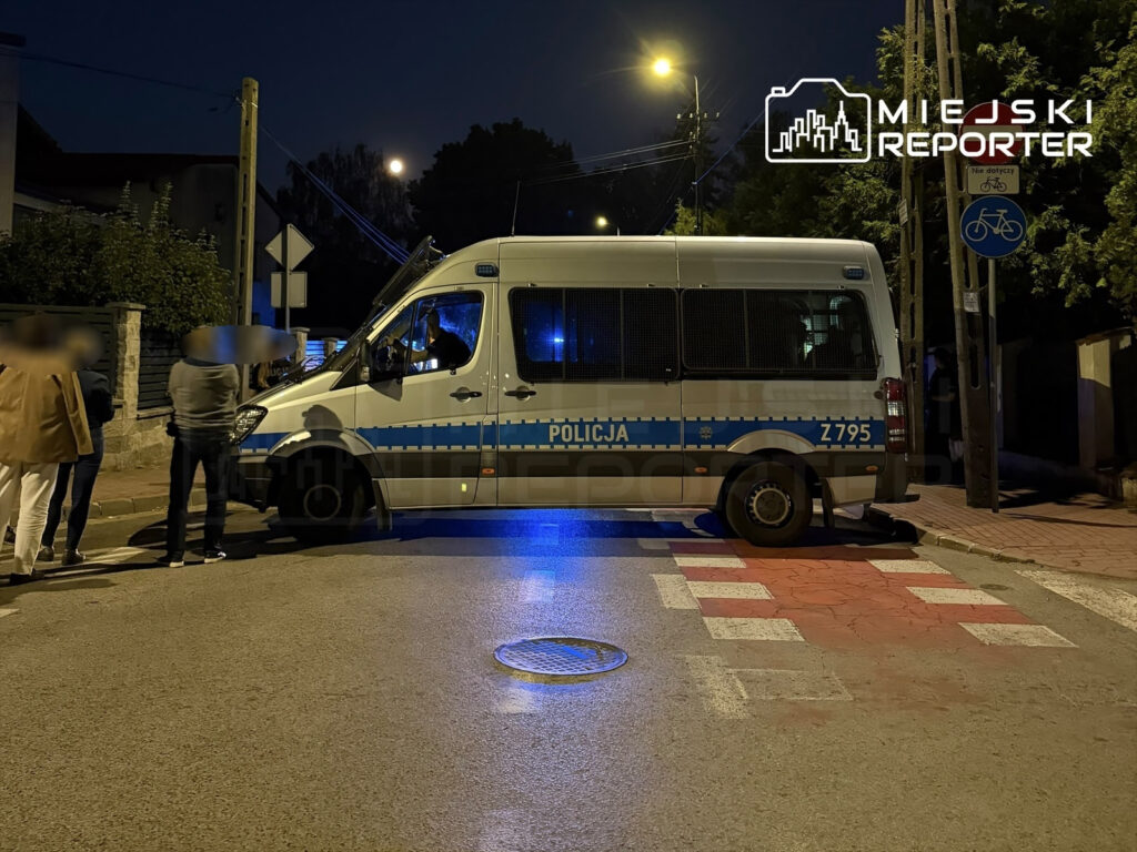 Funkcjonariusze policji stoją przy radiowozie na oświetlonej ulicy, podczas nocnej interwencji.