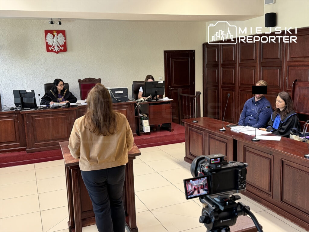 Kobieta w beżowej kurtce stoi przy mównicy, a sędzia w togach siedzi za biurkiem w sali sądowej.