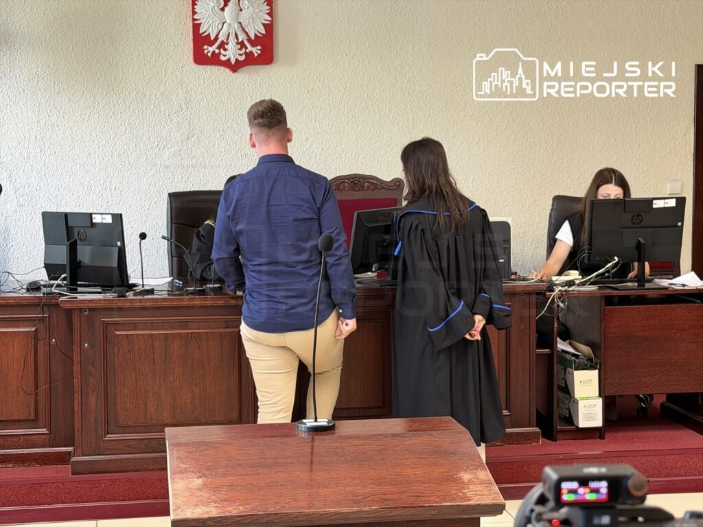 Mężczyzna w granatowej koszuli stoi przy mównicy, obok niego sędzia w togach, a w tle pracownik sądu przy komputerze.