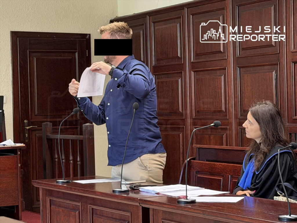 Mężczyzna w granatowej koszuli pokazuje dokumenty podczas rozprawy sądowej, siedząca obok kobieta w togach.