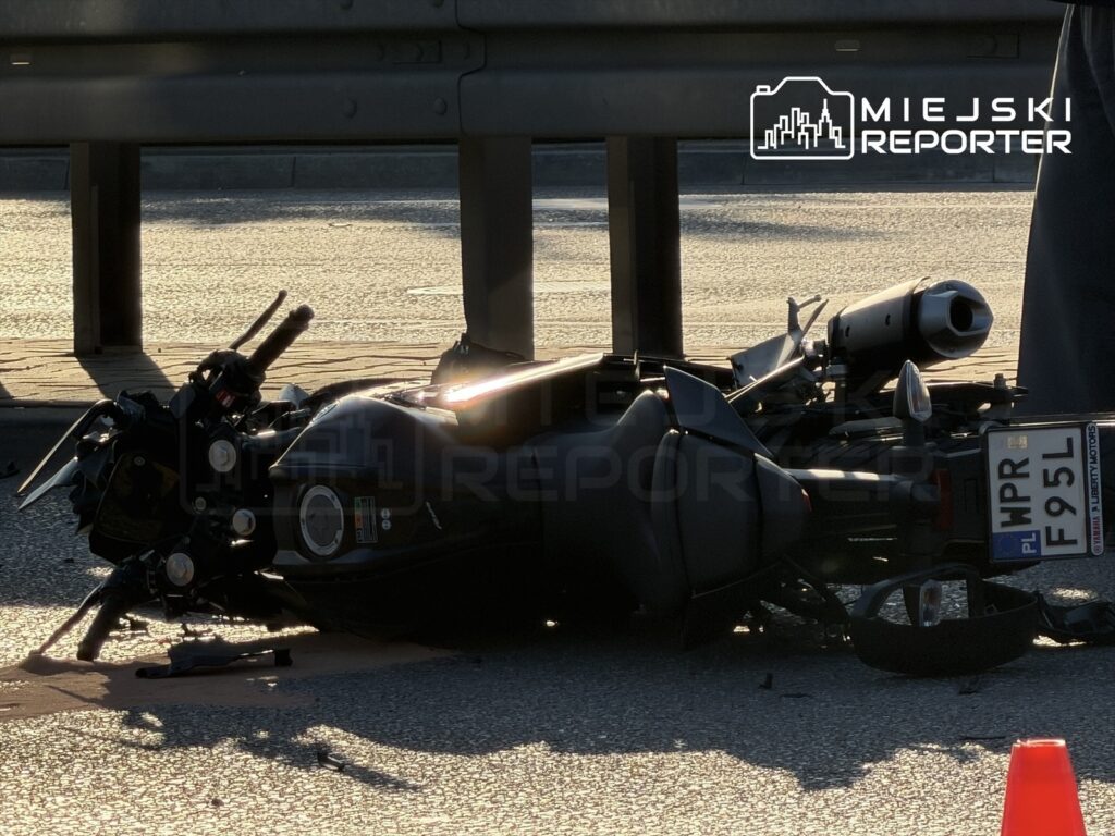 Motocykl leżący na jezdni, obok którego znajdują się elementy rozbitego pojazdu, w otoczeniu barierki ochronnej.