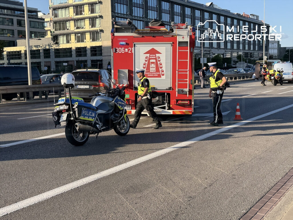 Policjanci w kamizelkach odblaskowych zabezpieczają miejsce wypadku na drodze, obok stoi wóz strażacki.
