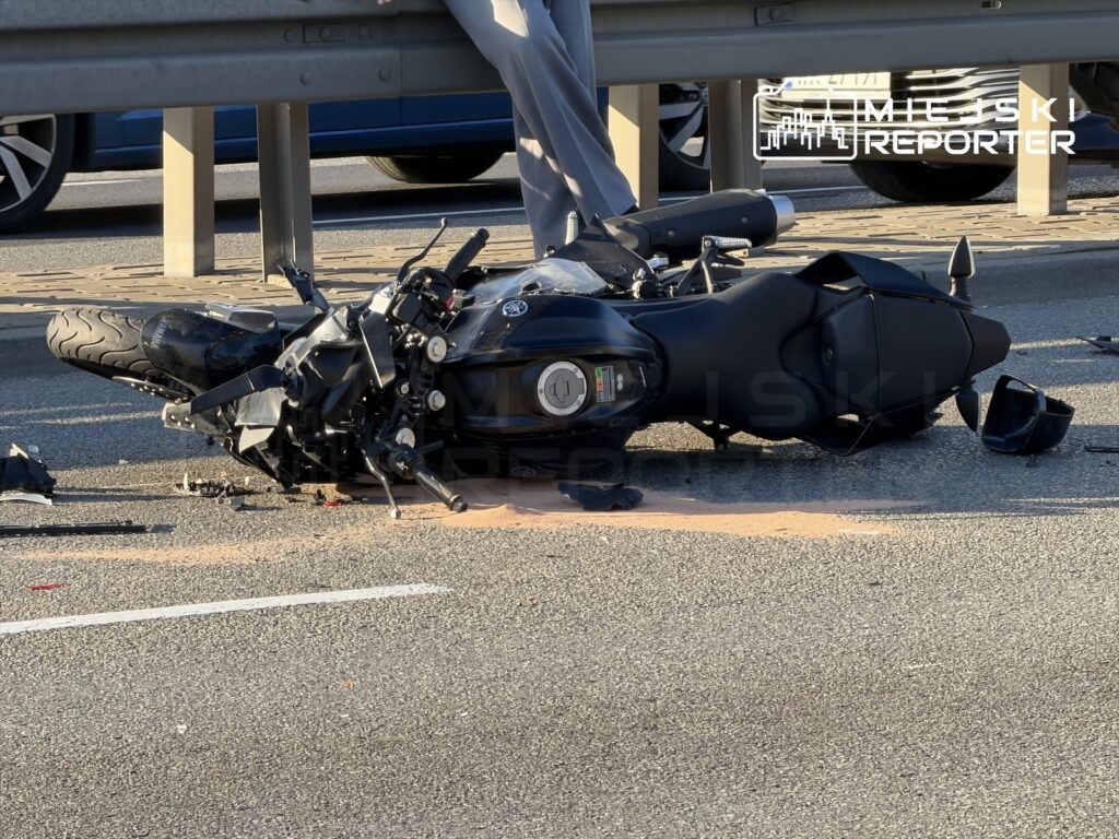 Motocykl leży na jezdni, obok widać uszkodzone elementy pojazdu, w tle ruch uliczny.