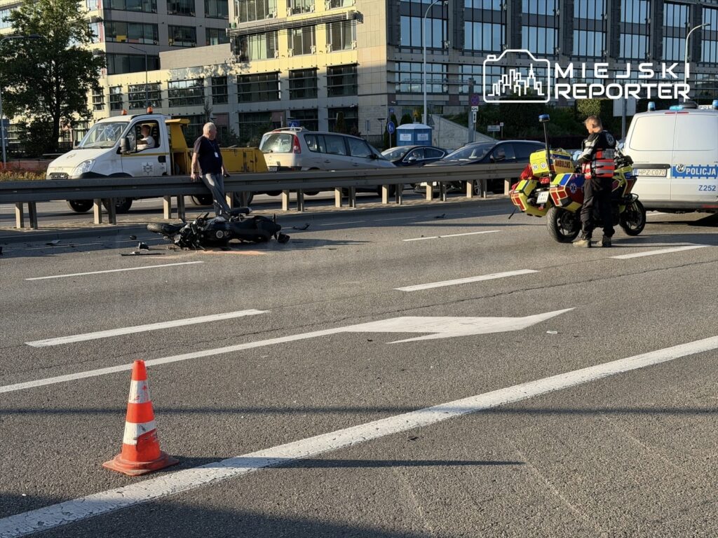 Motocyklista leży na jezdni obok przewróconego motocykla, a ratownik medyczny przybywa na miejsce wypadku.