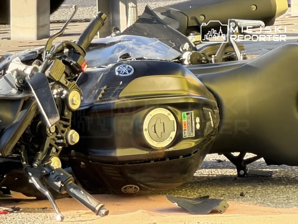 Motocykl leżący na boku na jezdni, z widocznym uszkodzonym elementem i włączonym światłem.