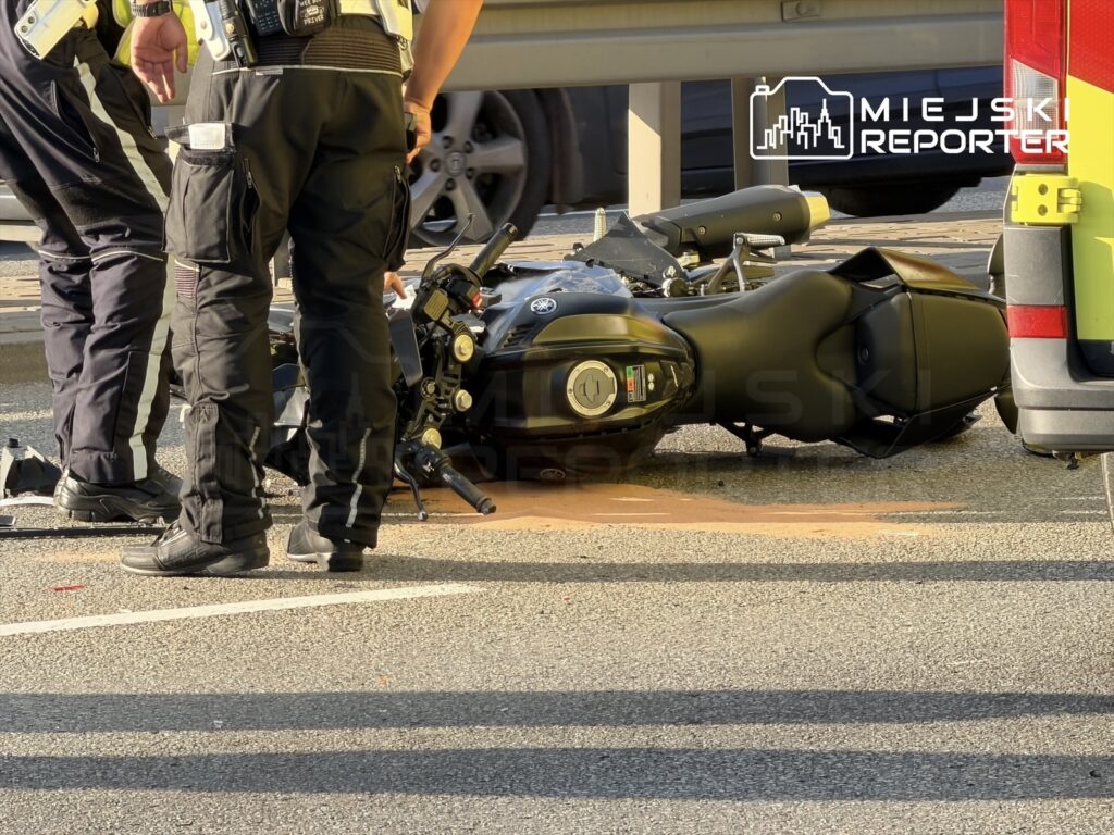 Motocyklista leży obok przewróconego motocykla, a policjanci badają miejsce zdarzenia na drodze.