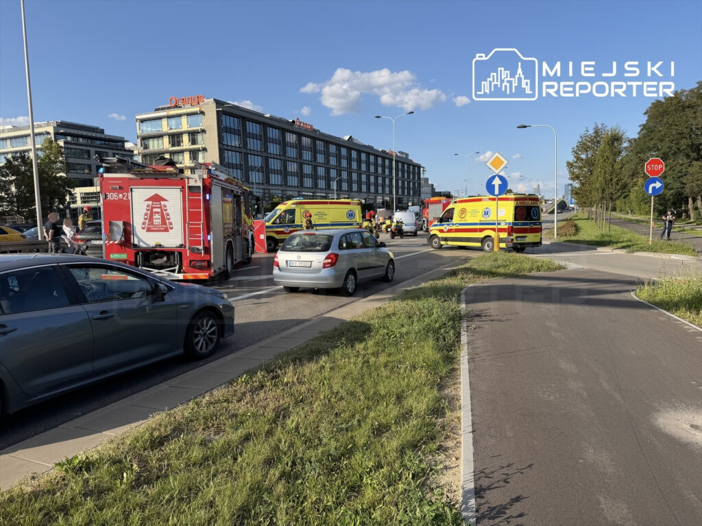 Na skrzyżowaniu zatrzymane są pojazdy ratunkowe, w tym straż pożarna i karetki, obok nowoczesnego biurowca.