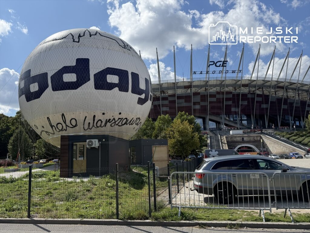 Balon widokowy z napisem "Hello Warszawa" stoi przed PGE Narodowym, otoczony zielenią i parkingiem.