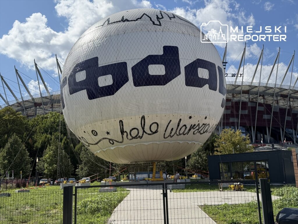 Balon z napisem "hello Warszawa" unosi się nad zielonym terenem w pobliżu stadionu, otoczony drzewami i ogrodzeniem.