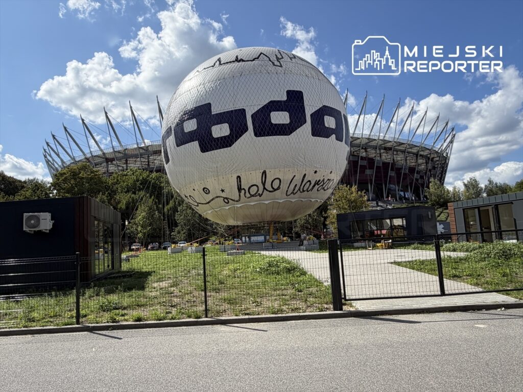 Balon z napisem "Hello Warsaw" unosi się nad zieloną przestrzenią, w tle nowoczesny stadion z charakterystycznymi daszkami.