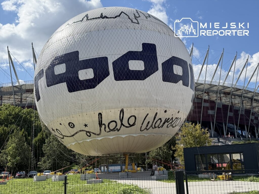 Balon widokowy z napisem "hello Warszawa" unosi się nad terenem przy nowoczesnym stadionie, w tle błękitne niebo.