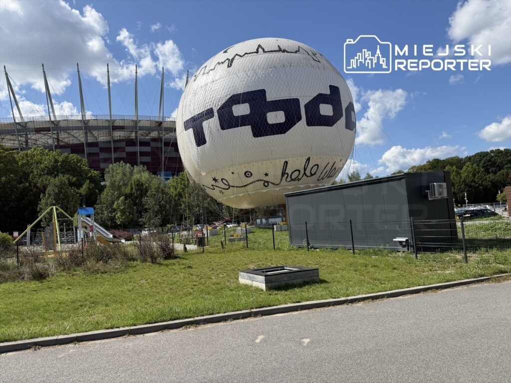 Balon widokowy unosi się nad terenem rekreacyjnym, w tle widoczny stadion z charakterystycznymi masztami.