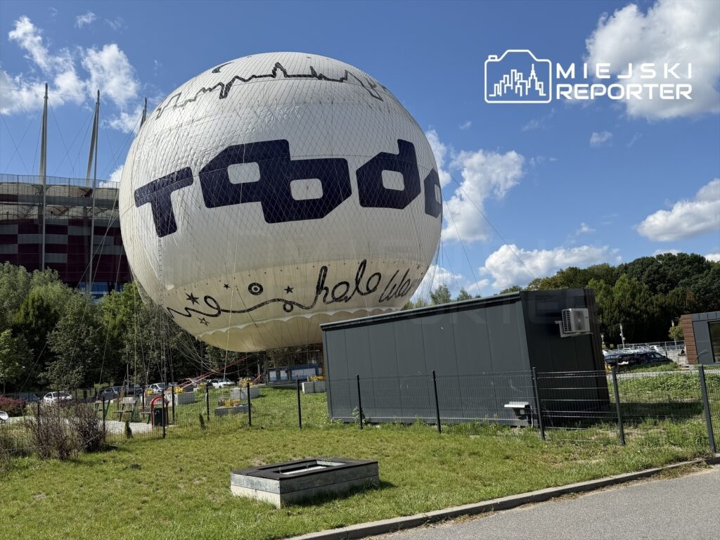 Balon wypełniony powietrzem stoi na trawie obok nowoczesnego budynku w otoczeniu zieleni i parkingu.