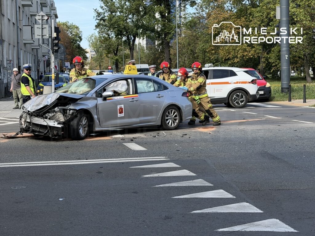 Ratownicy w hełmach pomagają w usuwaniu uszkodzonego samochodu z ruchliwej ulicy, obok stoją radiowozy.