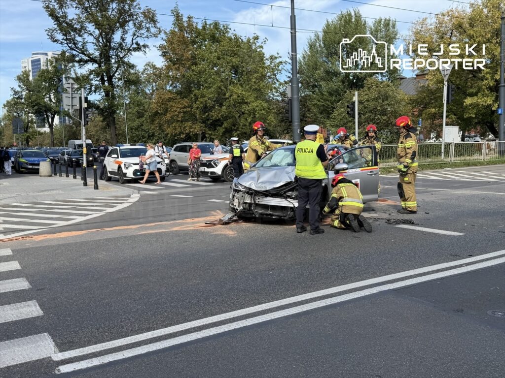 Na skrzyżowaniu dwóch ulic, strażacy oraz policjanci udzielają pomocy przy rozbitym samochodzie.