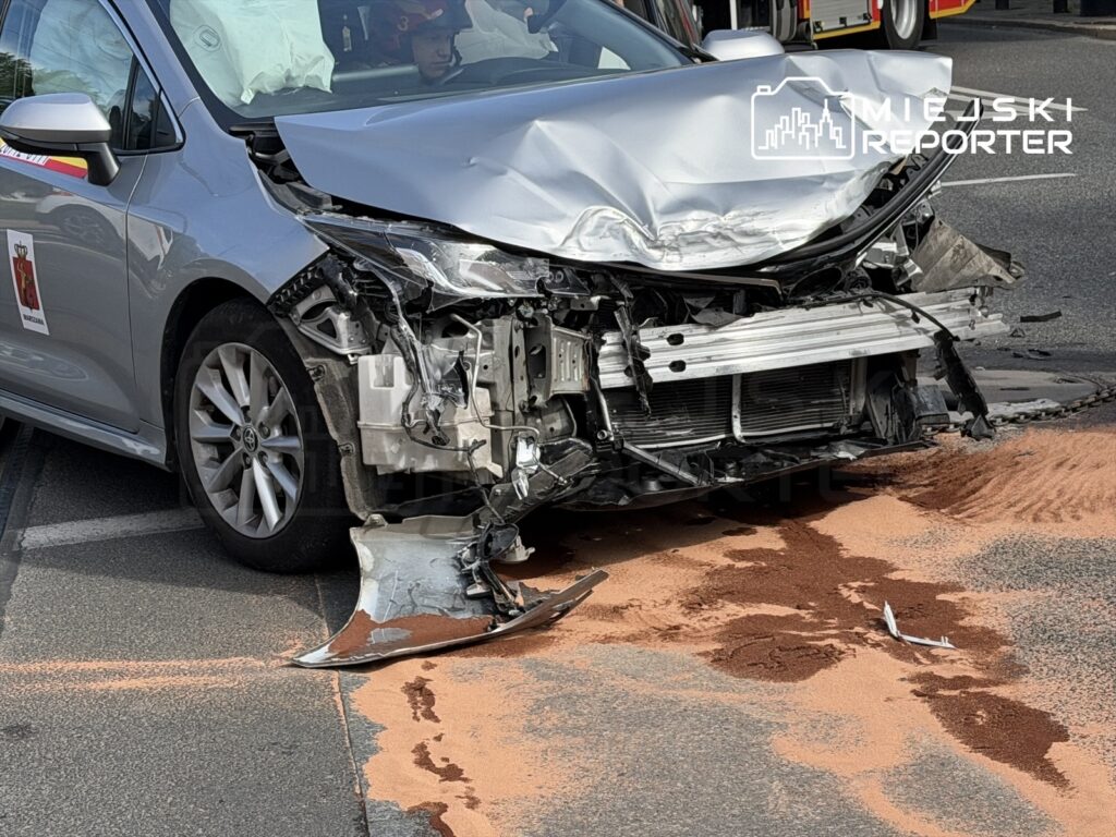 Rozbity samochód osobowy z uszkodzoną przednią częścią, obok rozrzucony proszek na jezdni. W tle widać straż pożarną.