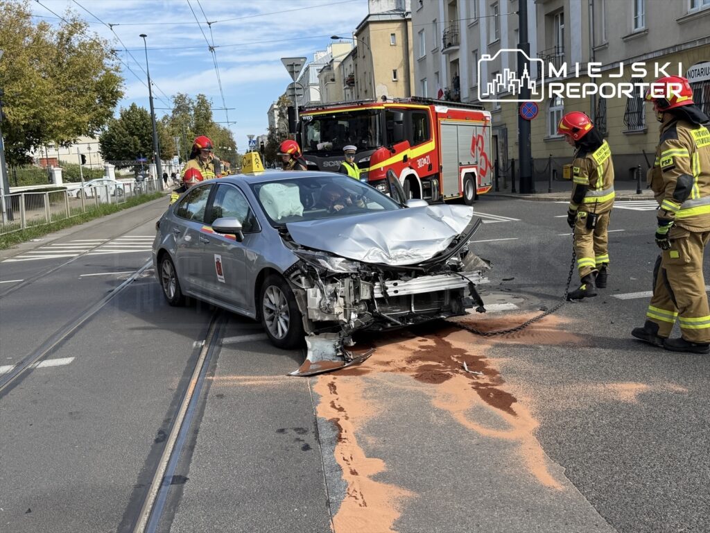 Rozbity samochód osobowy z uszkodzoną przednią częścią leży na torach tramwajowych, obok strażacy zabezpieczają miejsce zdarzenia.