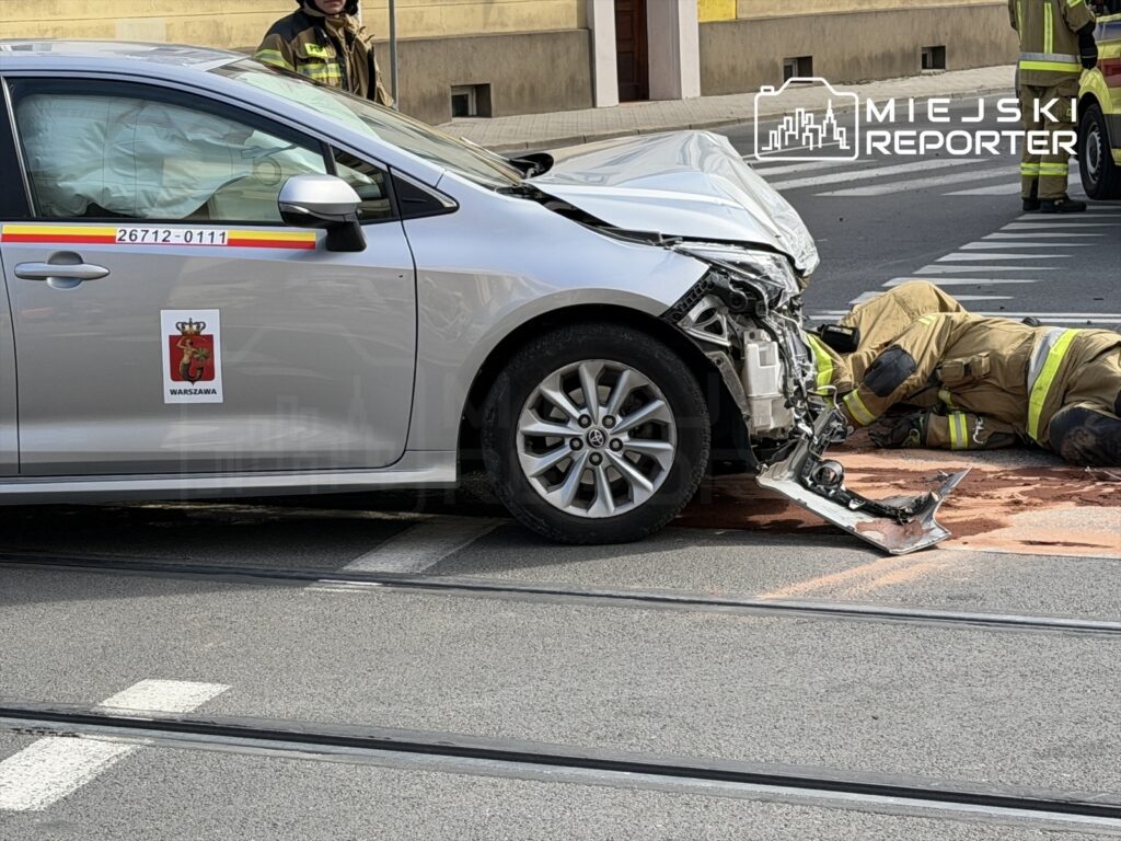 Uszkodzony samochód osobowy z widocznym uszkodzeniem przodu, obok leży strażak w kombinezonie ratunkowym, udzielający pomocy.