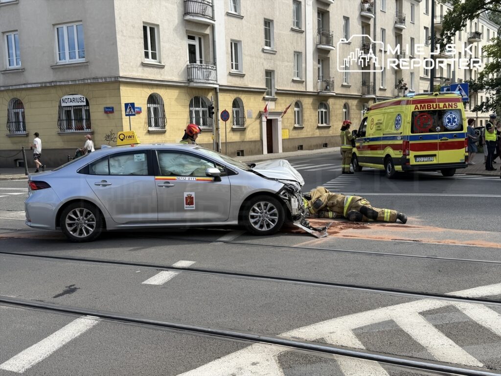 Złamany przód srebrnego samochodu osobowego, a obok leży strażak w mundurze, prowadzący działania ratunkowe na ulicy. W tle widoczne karetka pogotowia i przechodnie.