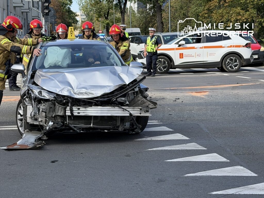 Grupa strażaków udziela pomocy kierowcy w uszkodzonym taksówce na skrzyżowaniu ulic.