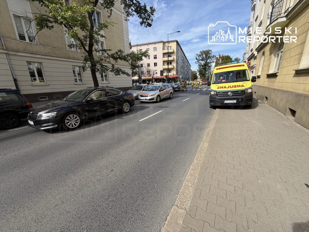 Na ulicy stoją rozbite samochody, w tym czarny pojazd z uszkodzeniem bocznym, obok parkuje karetka pogotowia.