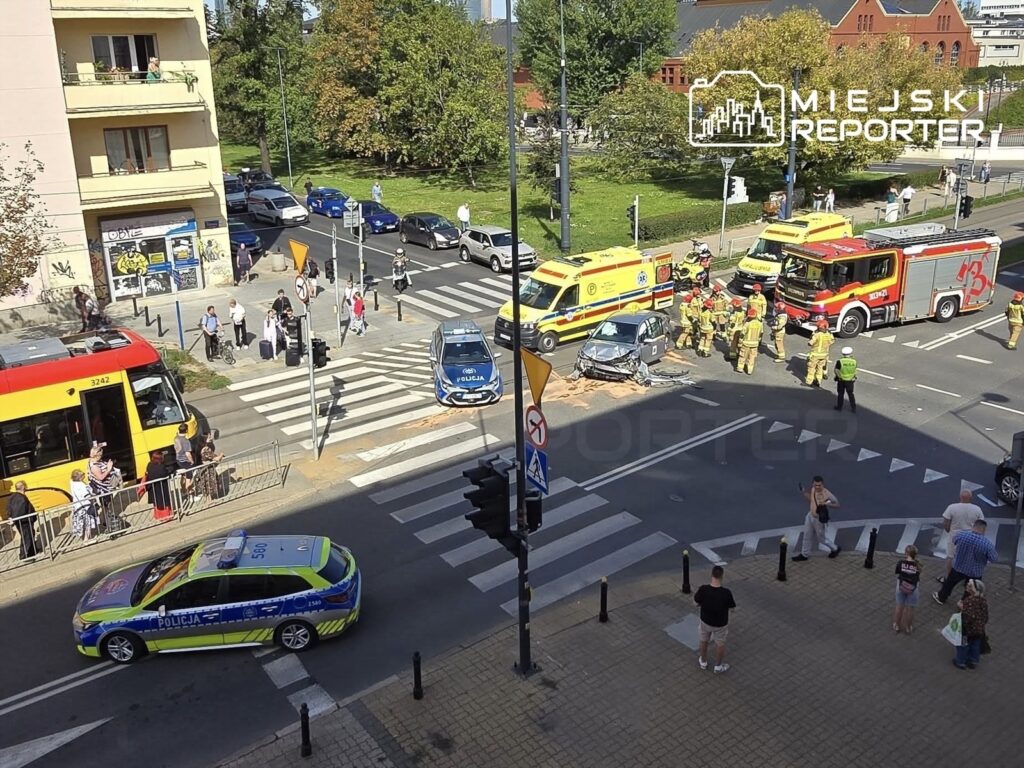 Na skrzyżowaniu stoją uszkodzone auta, a obok pracują strażacy i ratownicy medyczni. W tle widać tramwaj i przechodniów.