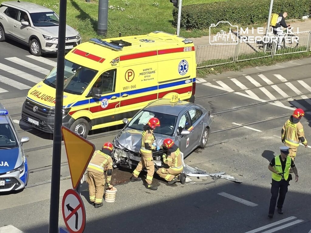 Ratownicy w hełmach badają uszkodzone auto na skrzyżowaniu, obok stoi ambulans i radiowóz policyjny.