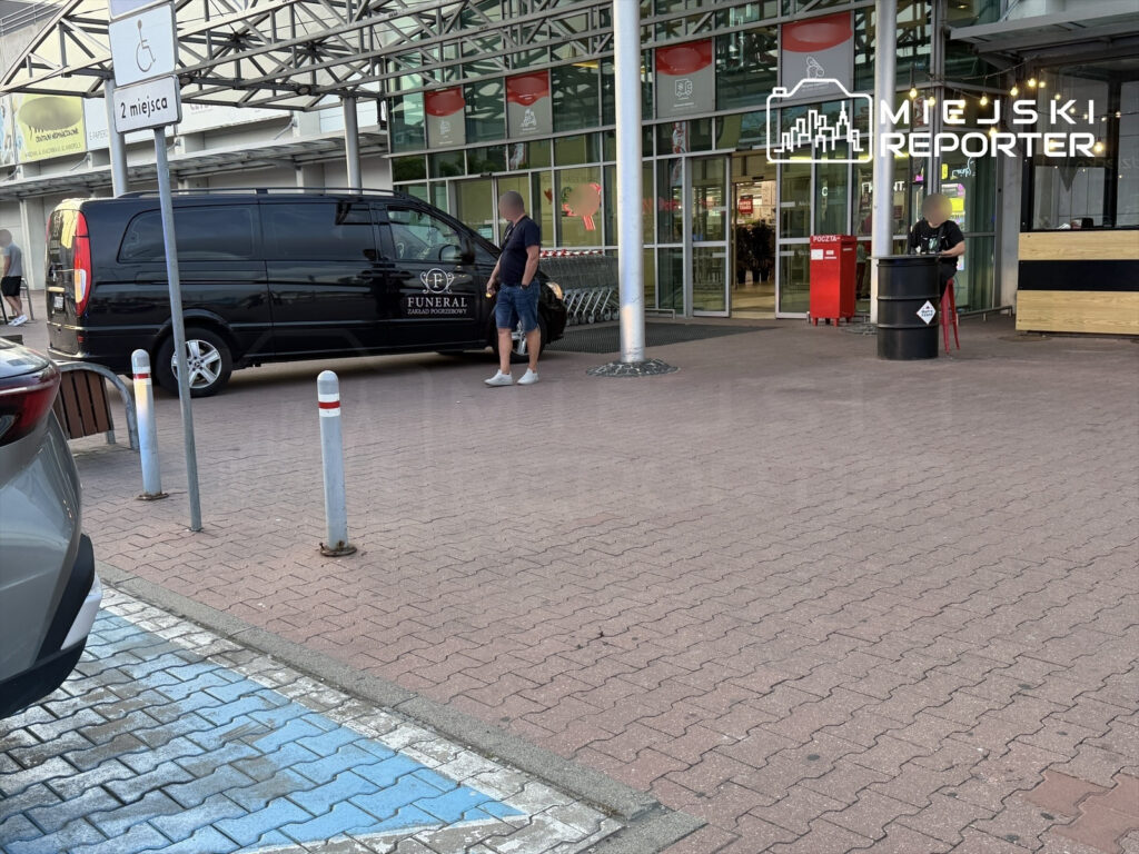 Mężczyzna rozmawia z pracownikiem przy czarnym samochodzie pogrzebowym przed wejściem do sklepu.