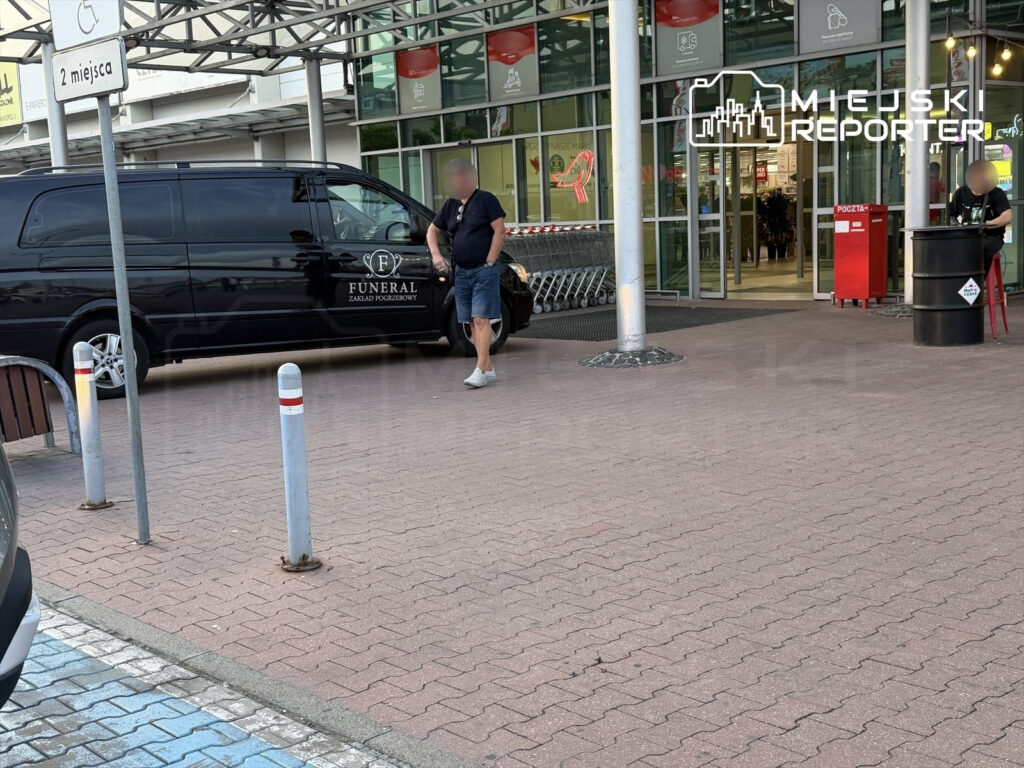 Mężczyzna w krótkich spodniach stoi obok czarnego samochodu pogrzebowego przed wejściem do sklepu.